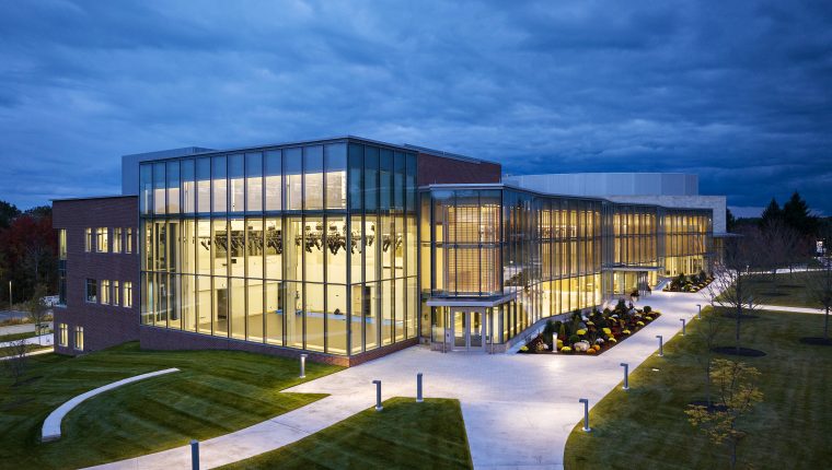 Modern building with glass facade illuminated interior surrounded by landscaped pathways and trees under a dusky sky