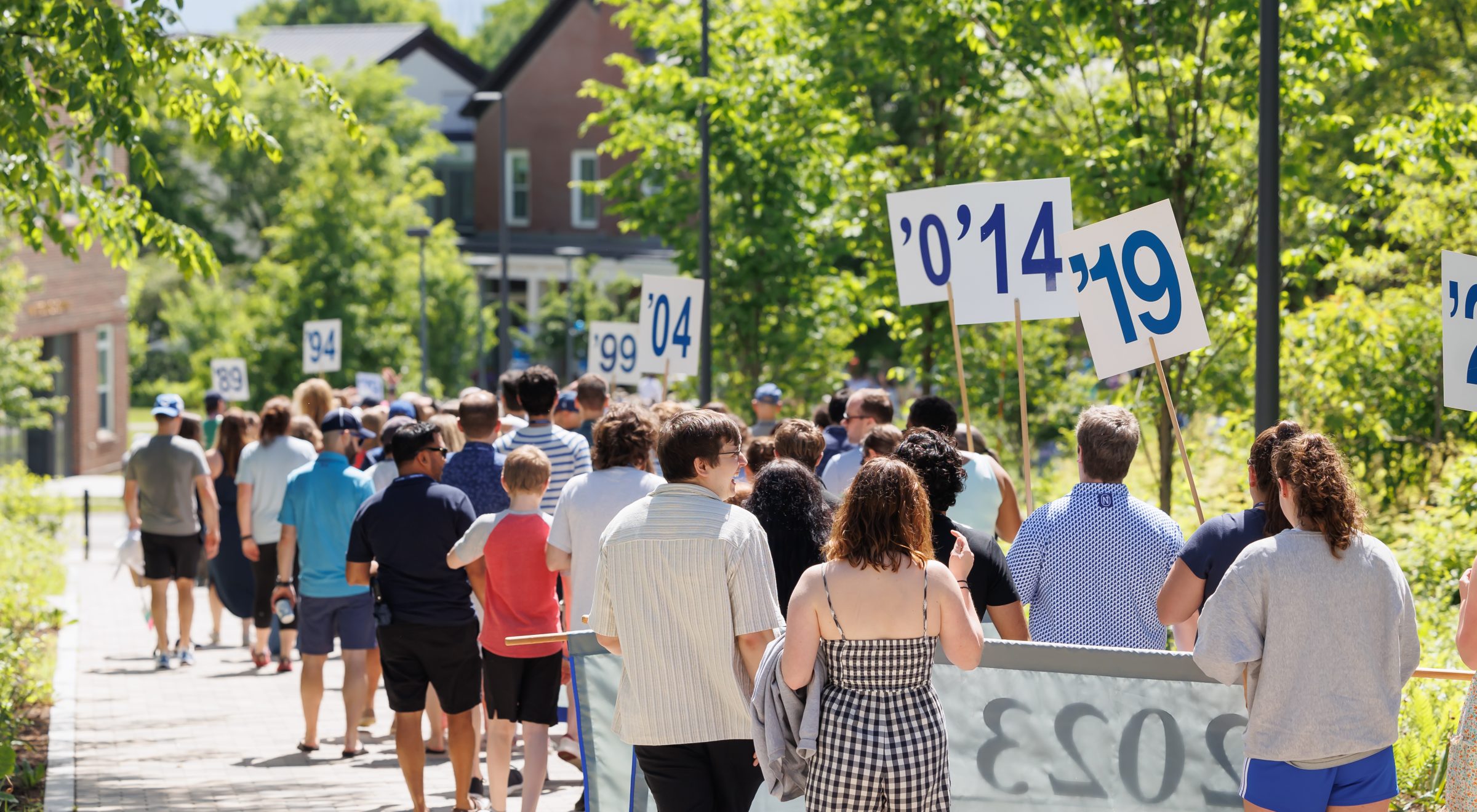 A crowd of people walking along a path holding signs with numbers under a sunny sky and surrounded by greenery